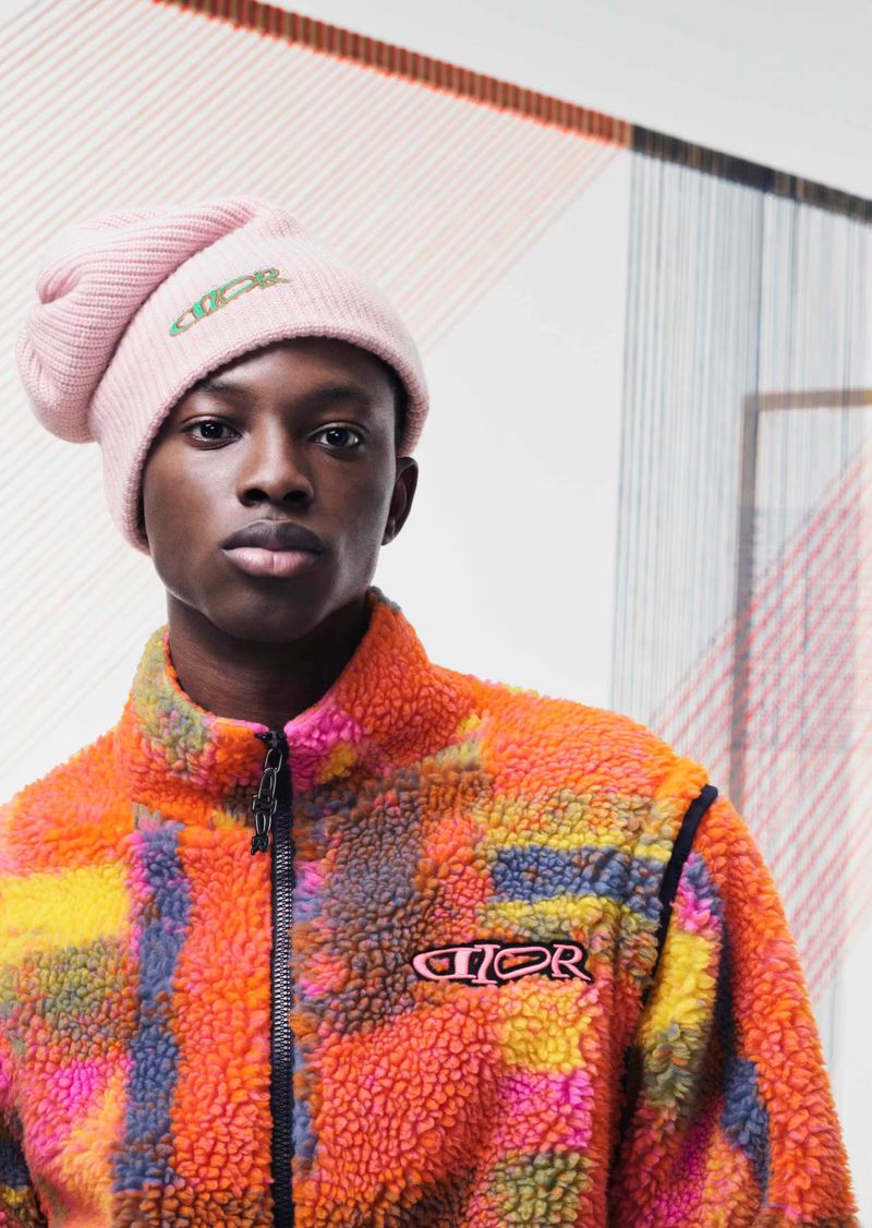 Young black male model in pink logo beanie and multicolored fleece zip jacket in a studio setting with an abstract art background.
