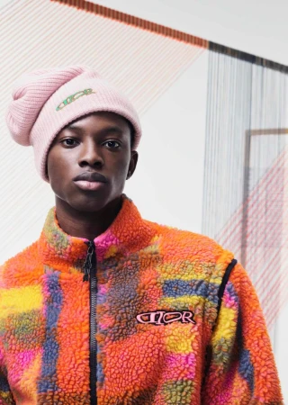 Young black male model in pink logo beanie and multicolored fleece zip jacket in a studio setting with an abstract art background.