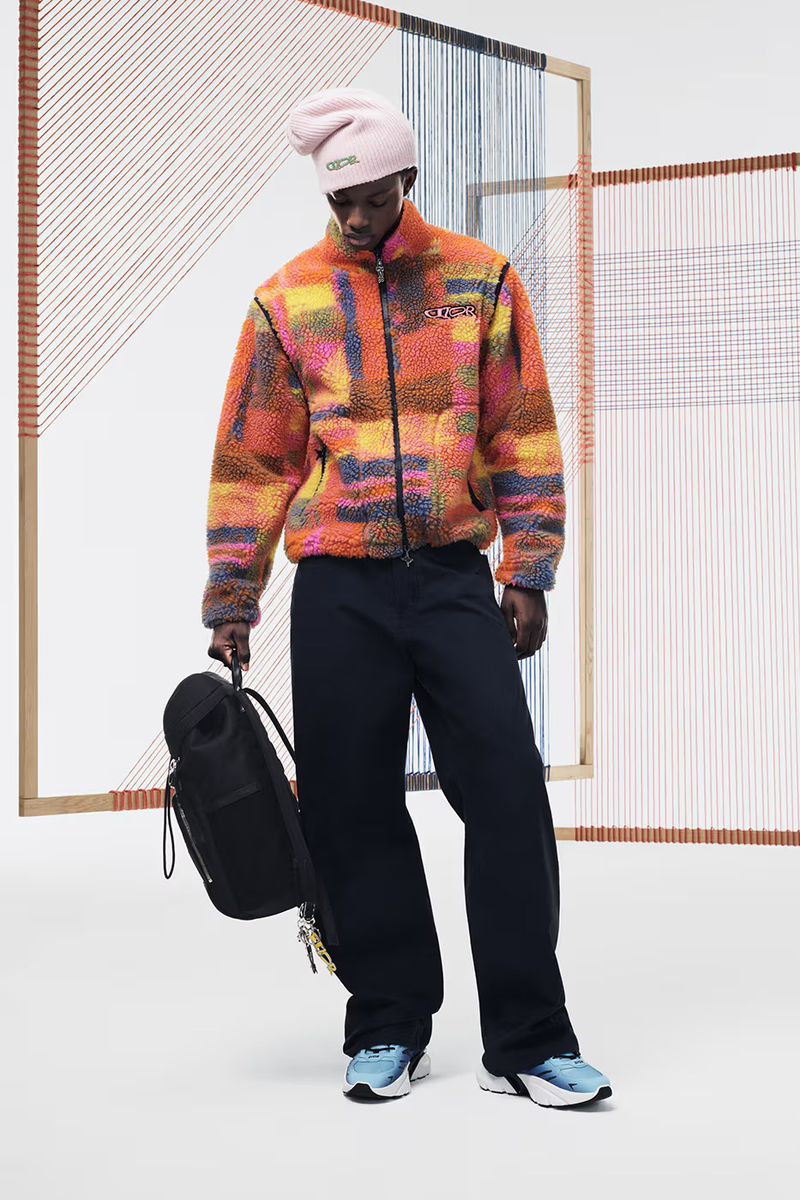 Male model in colorful jacket, black pants, beanie, and sneakers stands in a studio holding a black bag against an abstract backdrop.