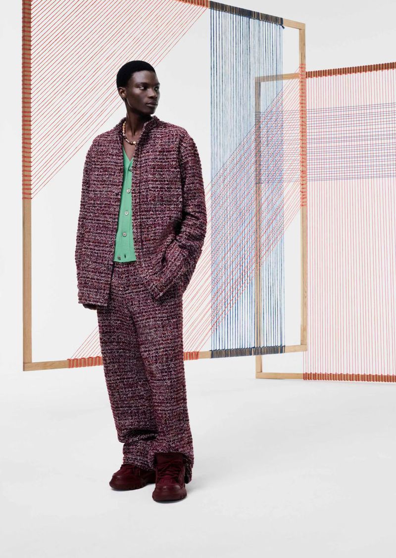 An African American male model stands in a photo studio wearing a burgundy tweed suit, posed against a backdrop of art installations.