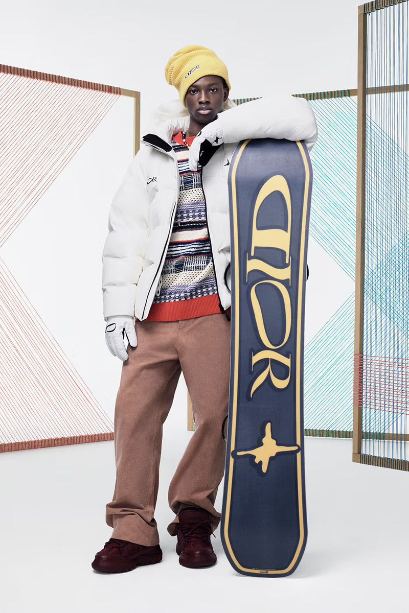 Man in winter fashion with white puffer jacket, knit sweater, snowboard, and beanie poses in a studio with geometric background.