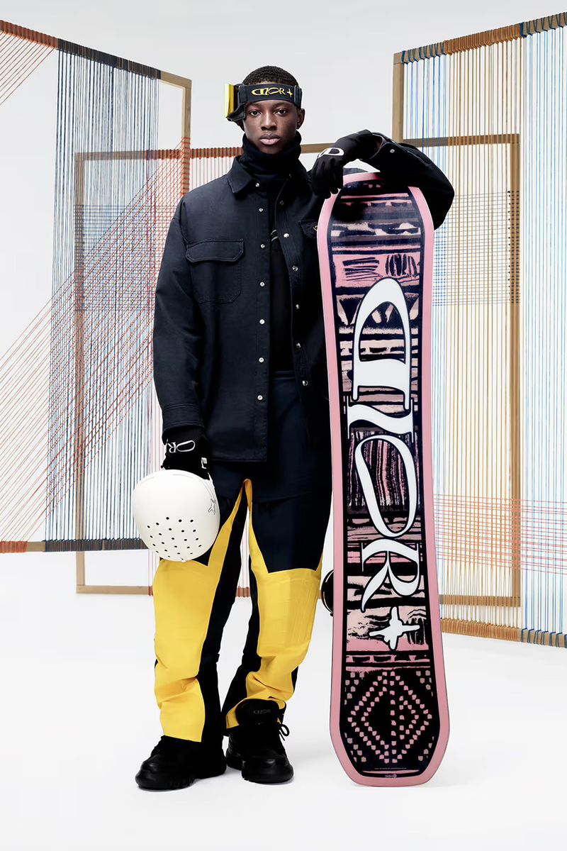 A male model stands with a snowboard, wearing a black jacket and yellow-accented pants, in a studio setting with geometric backdrop.