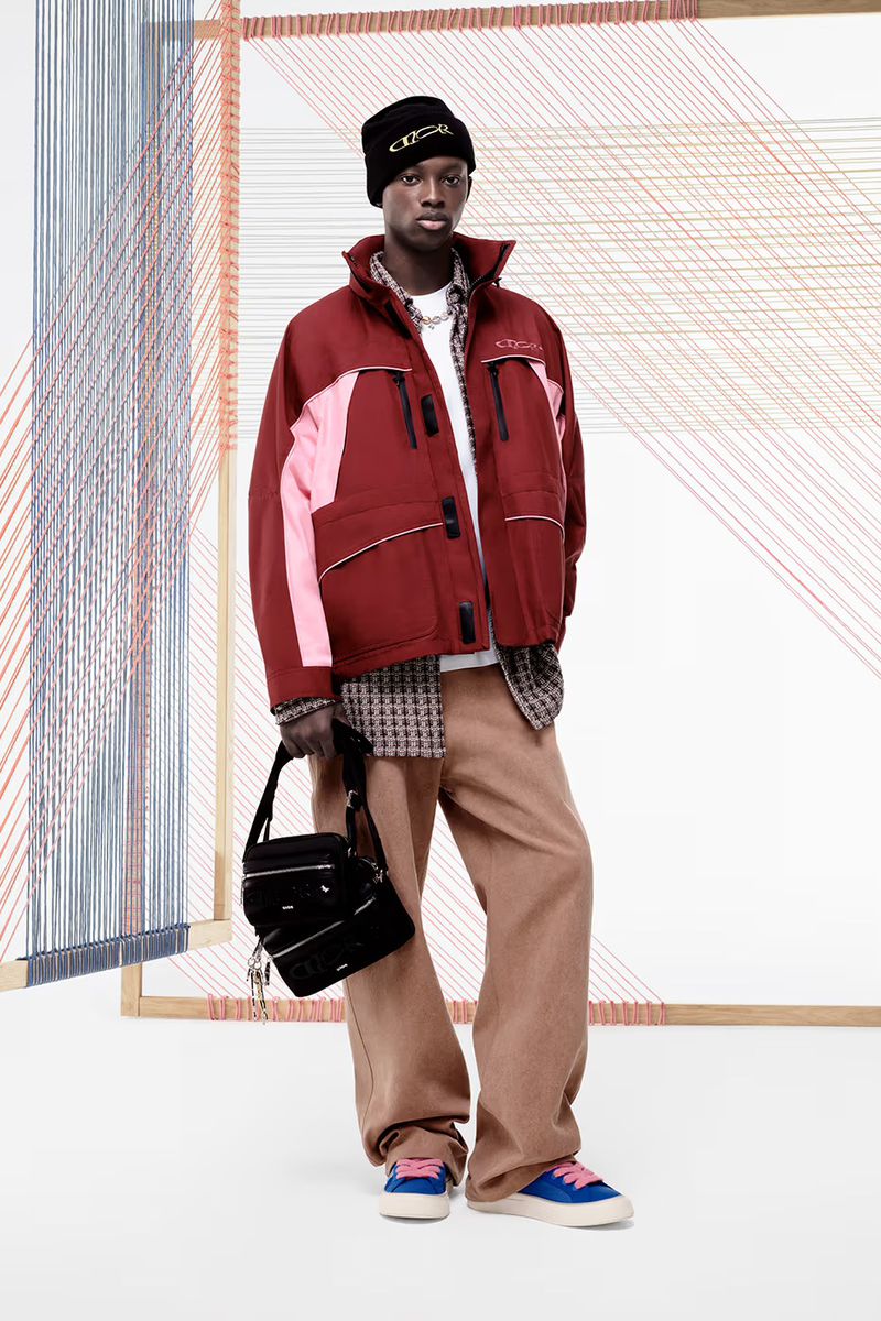 An adult male model poses in an outdoor studio wearing a maroon jacket, beige pants, a black beanie, and blue sneakers, carrying a bag.