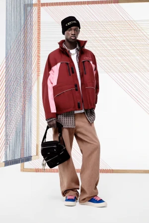 An adult male model poses in an outdoor studio wearing a maroon jacket, beige pants, a black beanie, and blue sneakers, carrying a bag.
