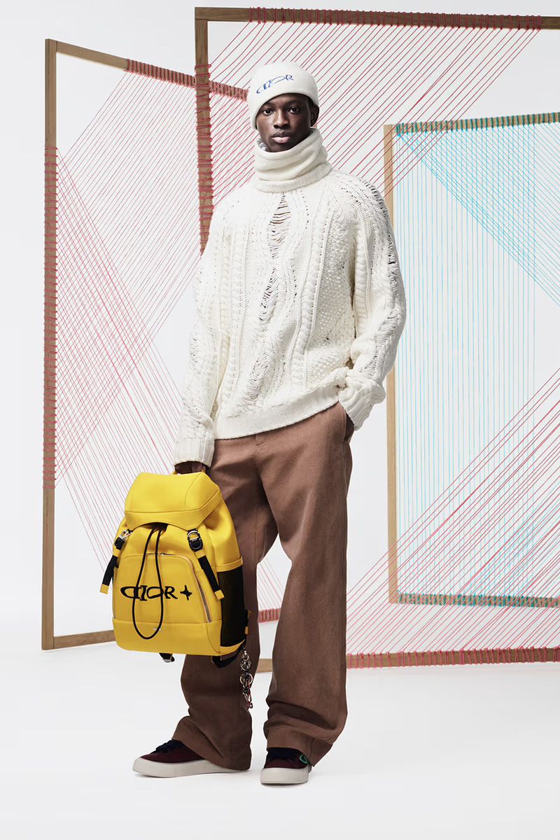 Male model in a cream cable-knit sweater, brown pants, and beanie posing with a yellow backpack against a geometric backdrop.