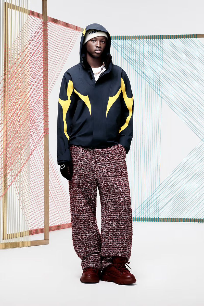 Full-length studio shot of a Black male model in a blue jacket with yellow accents, patterned pants, beanie, and red shoes against a white backdrop.