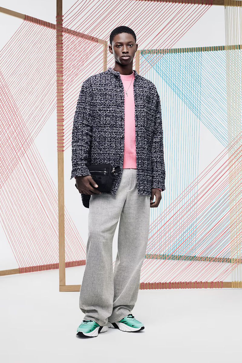 Male model in a studio setting wearing a pink sweater, tweed jacket, and gray pants, paired with colorful sneakers and a black bag.