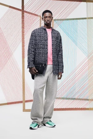 Male model in a studio setting wearing a pink sweater, tweed jacket, and gray pants, paired with colorful sneakers and a black bag.