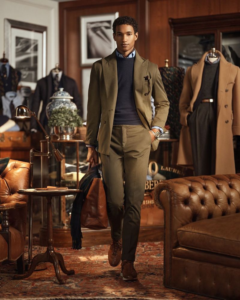 A dapper man in an olive suit walks confidently in a warm, wood-paneled room filled with classic furniture and tailored clothing.