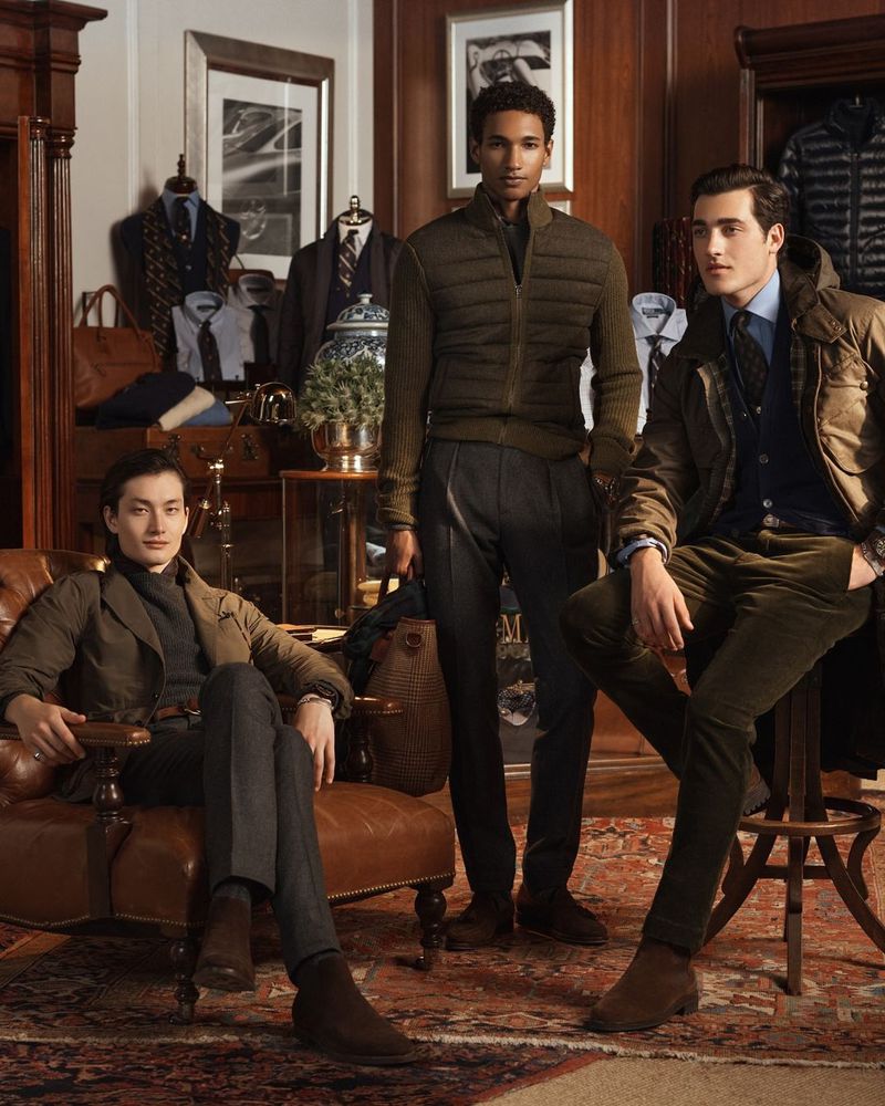 Three adult male models pose in a wood-paneled room, wearing casual fall fashion. A leather armchair and other props fill the scene.