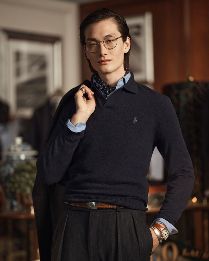 Portrait of a stylish Asian male wearing a blue sweater, grey trousers, and round glasses with a neutral facial expression.