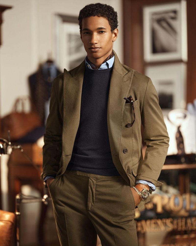 Dark-haired man poses indoors wearing an olive green blazer, sweater, and pants. He wears a watch and pocket square.