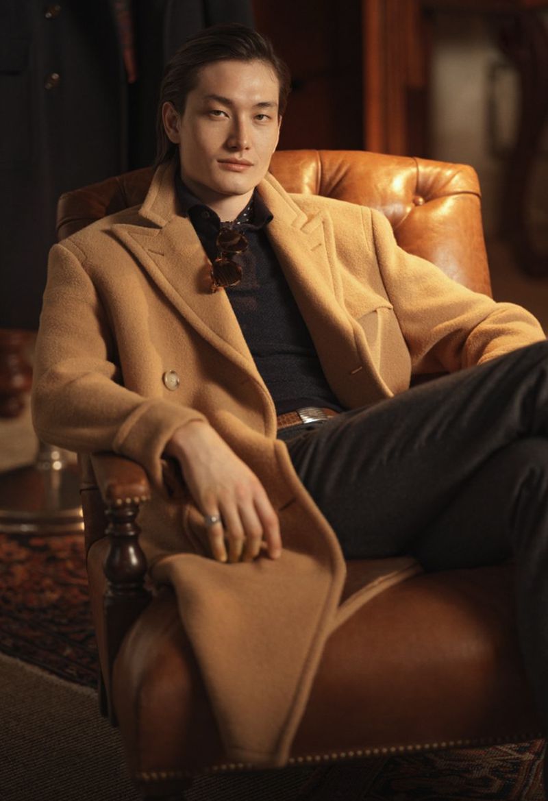 Portrait of a stylish adult male seated in a leather armchair, wearing a tan overcoat and dark pants in a warm-toned interior.