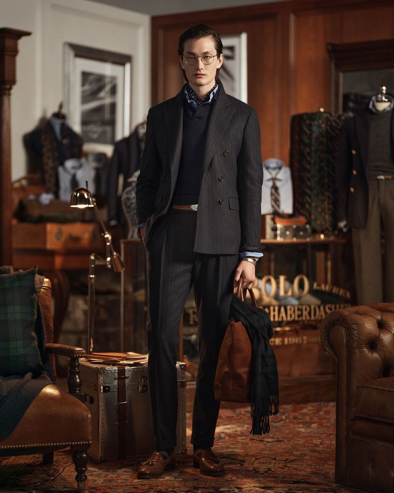 A man models a pinstripe suit in an indoor setting, posing in front of clothing displays, vintage luggage, and a leather armchair.