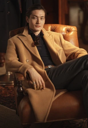 East Asian male model seated in a leather armchair wearing a camel wool coat and dark shirt for Polo Ralph Lauren.