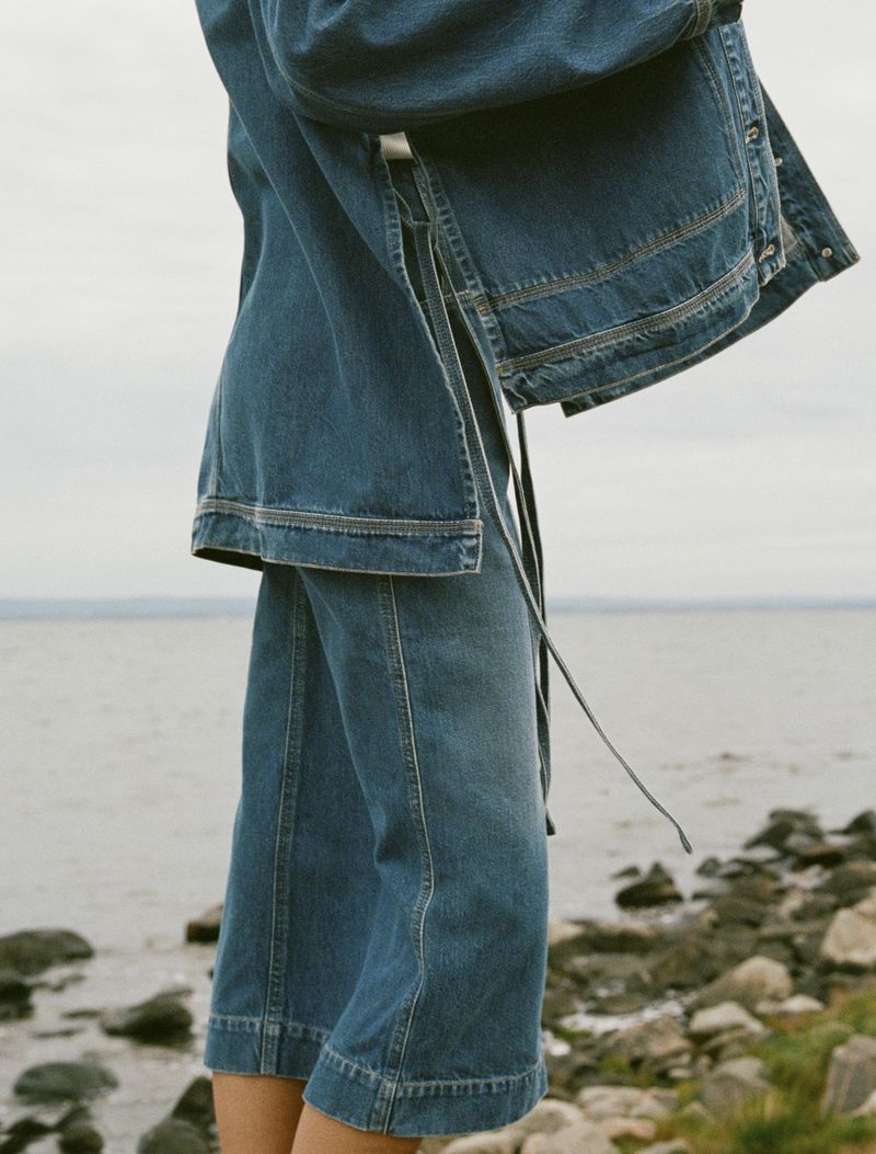 Mid-section view of a person wearing a blue denim jacket and capri pants standing outdoors near a body of water.