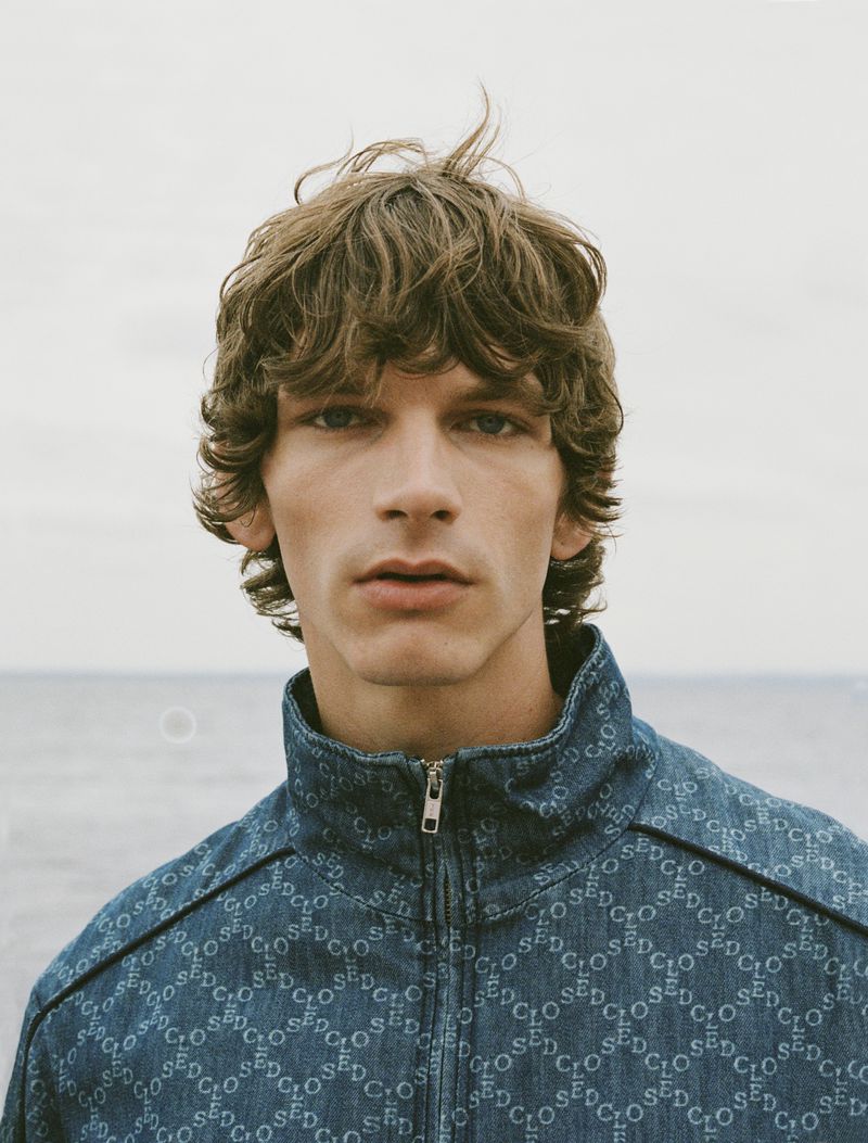 Close-up shot of a young adult man with curly brown hair wearing a blue patterned jacket against a soft sky and water background.