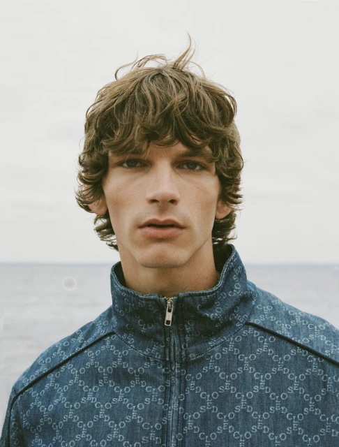 Close-up shot of a young adult man with curly brown hair wearing a blue patterned jacket against a soft sky and water background.