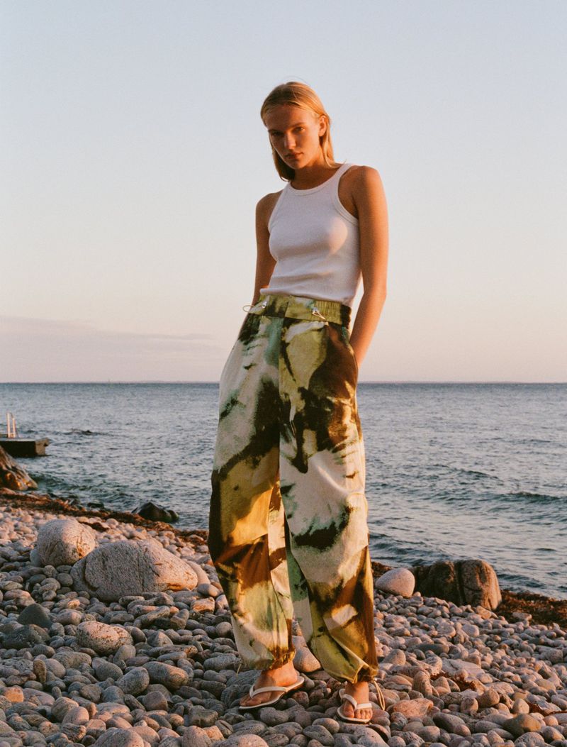 Blond woman poses on a rocky beach in stylish patterned pants and a white tank top, with the sea and sky in the soft background.