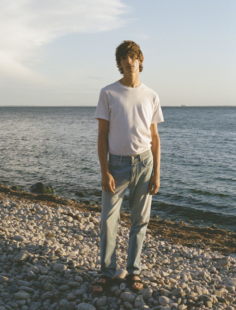 A young man stands on a rocky beach in jeans and a white t-shirt, facing the camera, with the ocean and sky in the background.