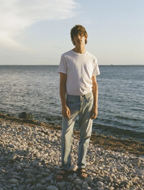 A young man stands on a rocky beach in jeans and a white t-shirt, facing the camera, with the ocean and sky in the background.