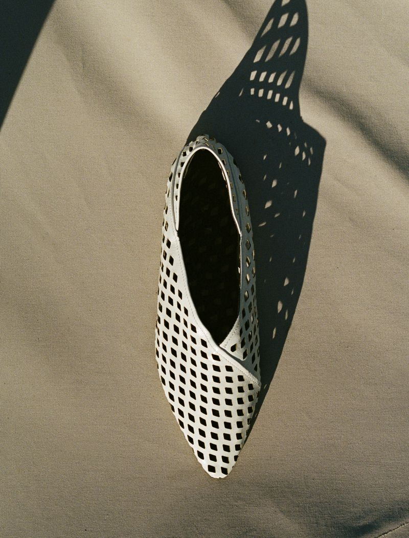 White leather open-weave ballet flat with pointy toe casts a sharp shadow on a light taupe fabric surface in simple studio shot.