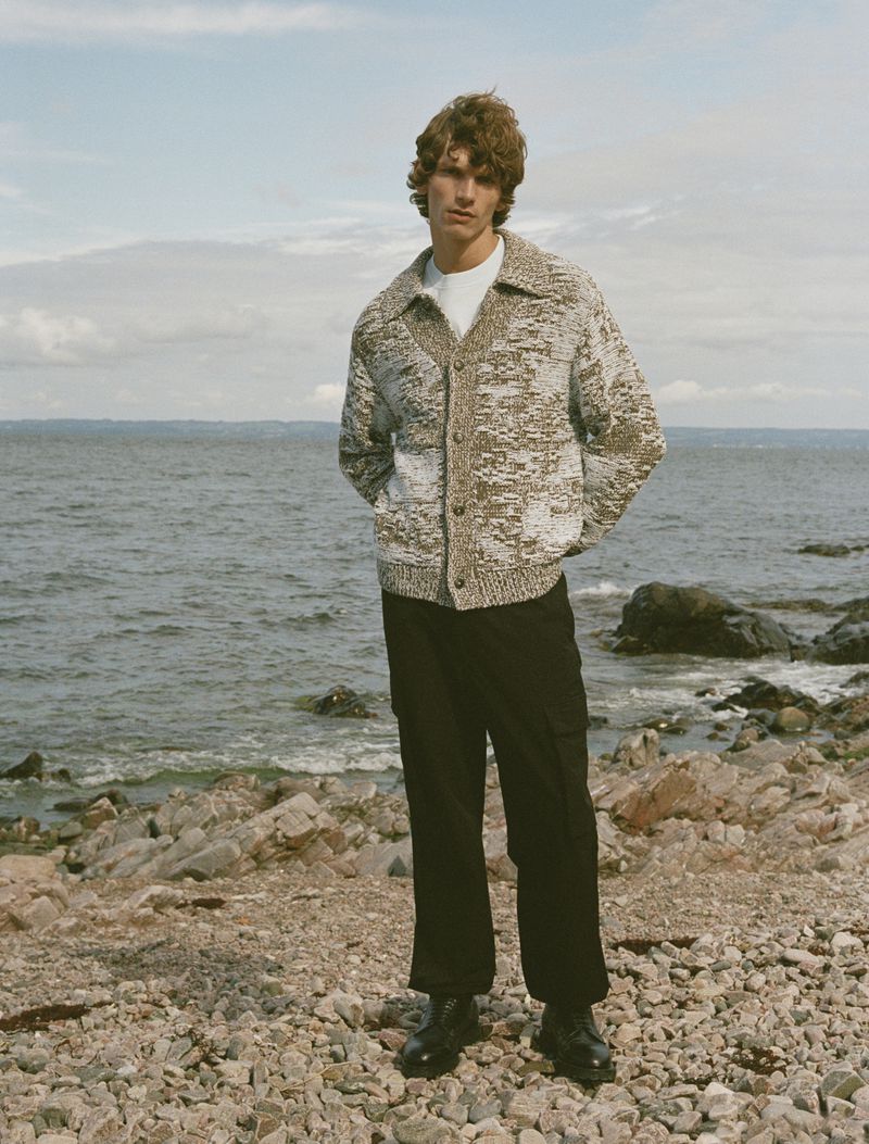 A fair-skinned adult male stands on a rocky beach wearing a buttoned speckled cardigan, dark pants, and black dress shoes, with the ocean backdrop.