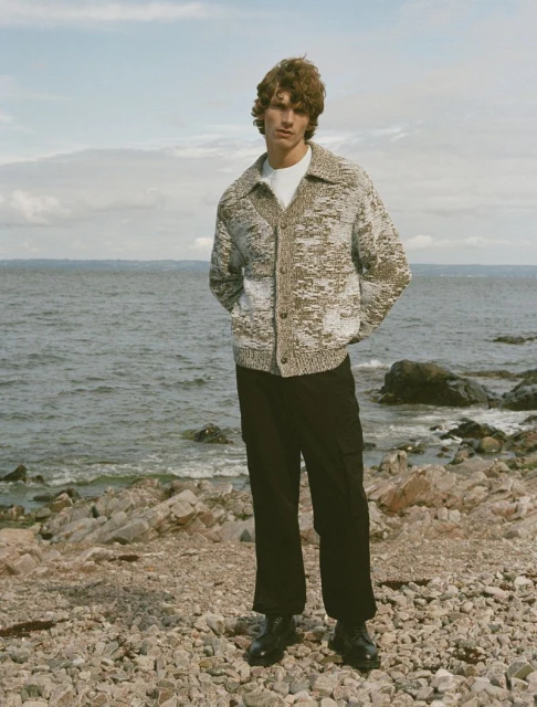 A fair-skinned adult male stands on a rocky beach wearing a buttoned speckled cardigan, dark pants, and black dress shoes, with the ocean backdrop.