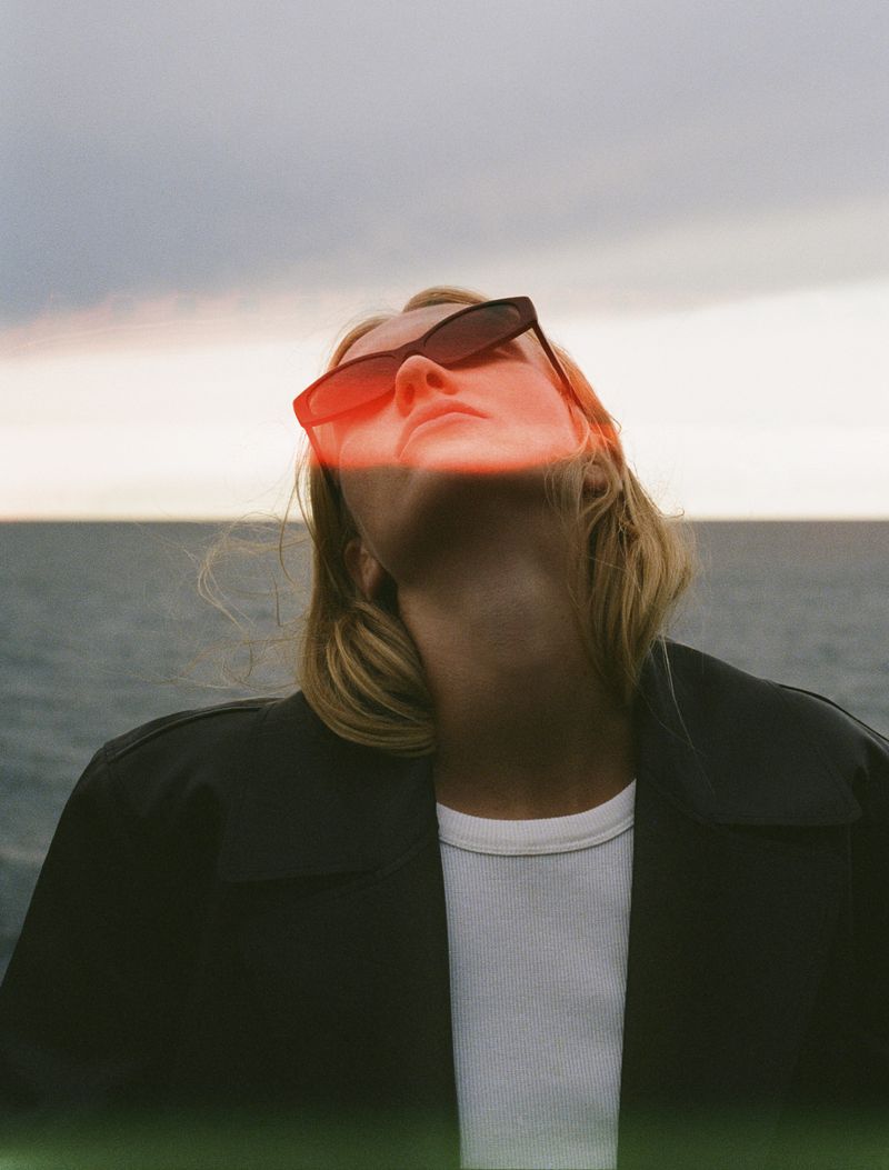Adult female gazes skyward in a low-angle portrait. She is wearing sunglasses and a jacket against a backdrop of ocean and cloudy sky.