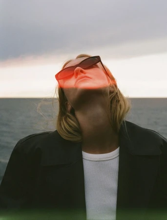 Woman looking upward wearing dark sunglasses, a black jacket, and a white t-shirt against a dusk ocean horizon with red light leak.