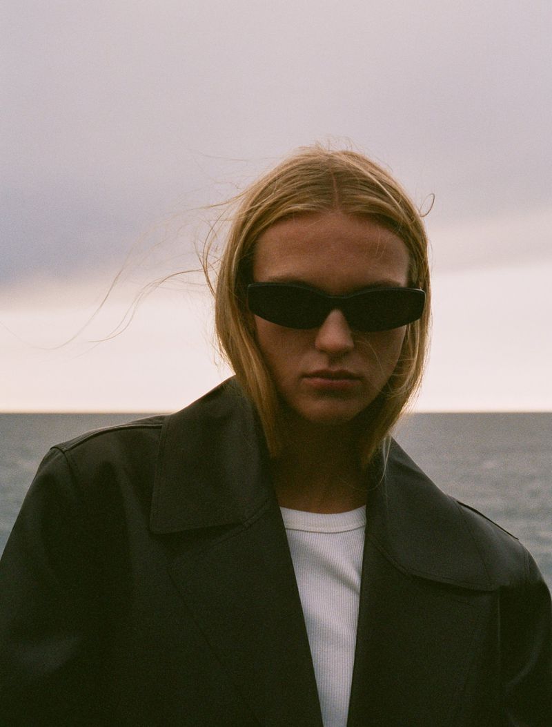 A woman wearing sunglasses and a trench coat stands outdoors with a cloudy sky and ocean in the background, shot in portrait orientation.