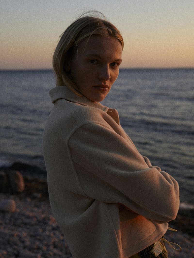 Woman in casual jacket poses on a rocky beach as the sun sets over a tranquil ocean, creating a serene and contemplative atmosphere.