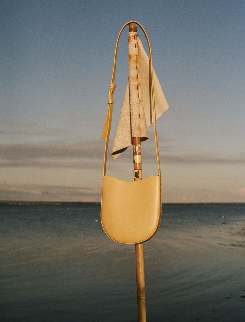 A yellow leather purse is hanging from a pole on a beach with blue sky and water in the background, creating a minimalist, modern aesthetic.
