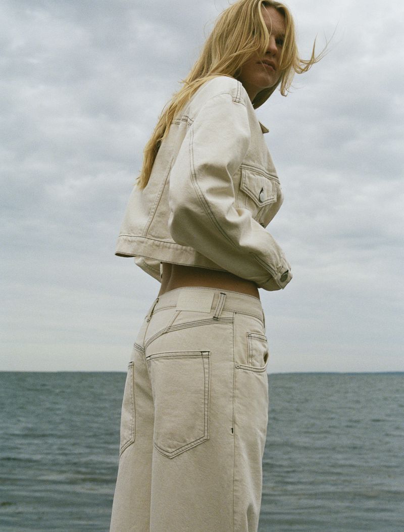 A woman with blonde hair poses in a crop top jacket and wide leg pants in an outdoor scene with a cloudy sky and a body of water.