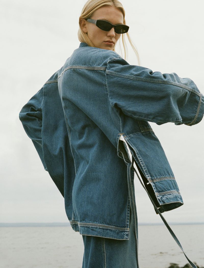 A blonde woman with sunglasses wears a denim jacket and pants in a natural light outdoor setting with a hazy sky and body of water.