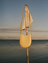 A yellow leather shoulder bag hangs from a wooden pole against a calm coastal water horizon under a warm evening sky.
