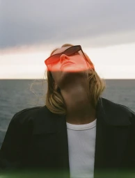Woman looking upward wearing dark sunglasses, a black jacket, and a white t-shirt against a dusk ocean horizon with red light leak.