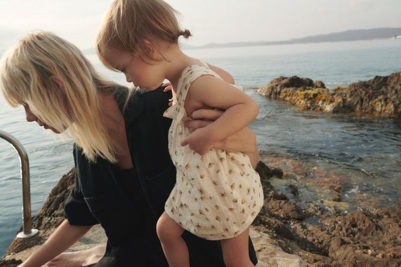 Woman and child by the sea, standing on a rocky outcrop, with the child in a floral romper and the woman in a dark dress.
