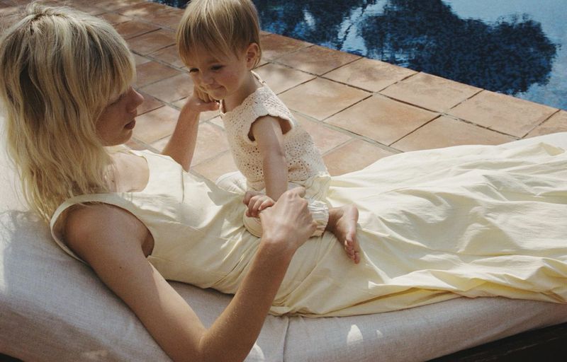 A woman and a toddler lounge on an outdoor chaise near a pool, bathed in soft, diffused daylight. The scene exudes warmth and relaxation.