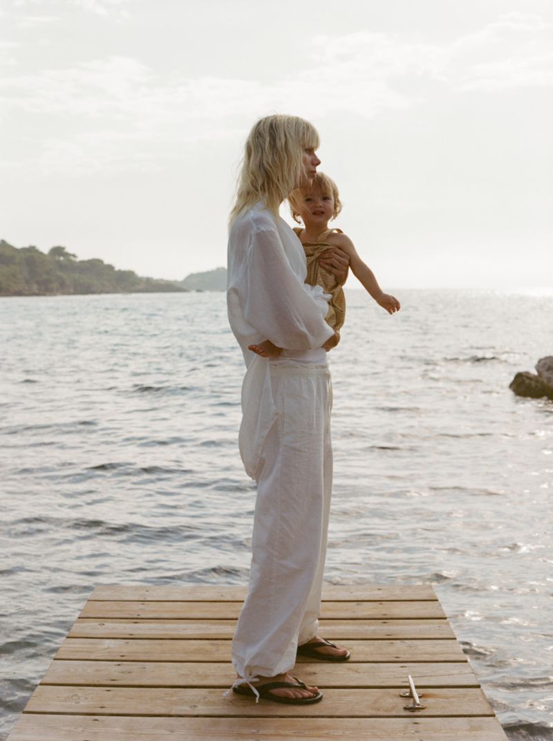 A blonde woman in white holds a baby in front of the ocean on a wooden dock on a cloudy day in a medium shot.