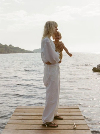 A blonde woman in white holds a baby in front of the ocean on a wooden dock on a cloudy day in a medium shot.