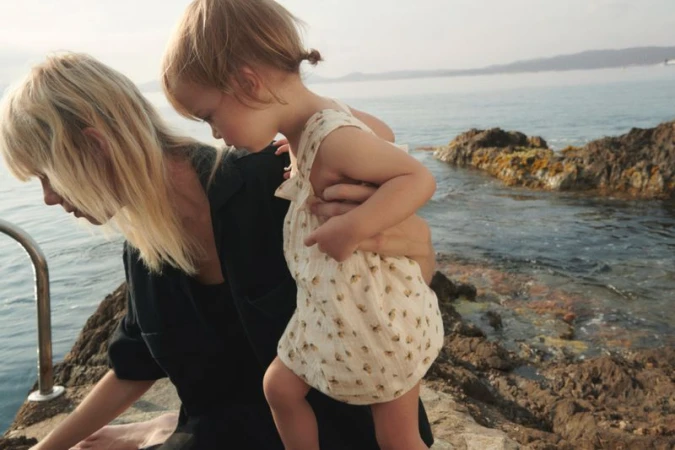 Marjan Jonkman holding a toddler in a patterned light romper against a rocky seashore for the Zara Sea Walk campaign.