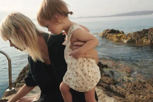Marjan Jonkman holding a toddler in a patterned light romper against a rocky seashore for the Zara Sea Walk campaign.