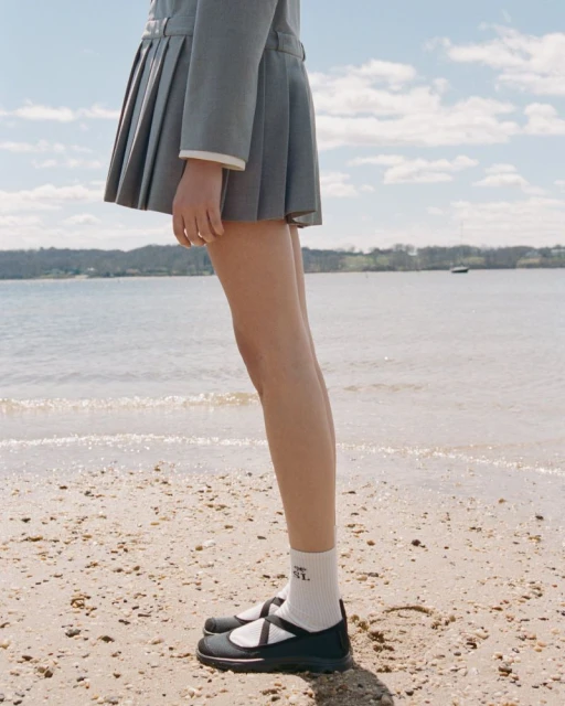 Young woman on a beach wearing a gray pleated skirt, socks, and black Mary Jane shoes, with an ocean and cloudy sky background.