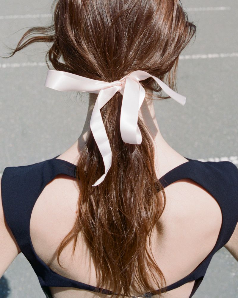Woman with brown hair styled in a low ponytail and tied with a pink satin ribbon bow, wearing a black halter top, seen from the back.