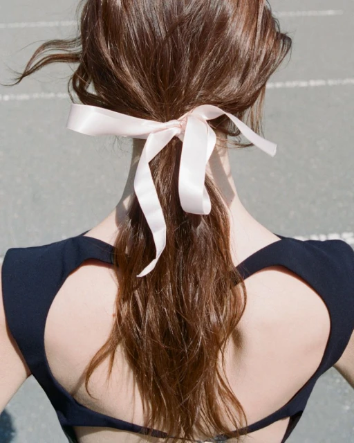 Woman with brown hair styled in a low ponytail and tied with a pink satin ribbon bow, wearing a black halter top, seen from the back.