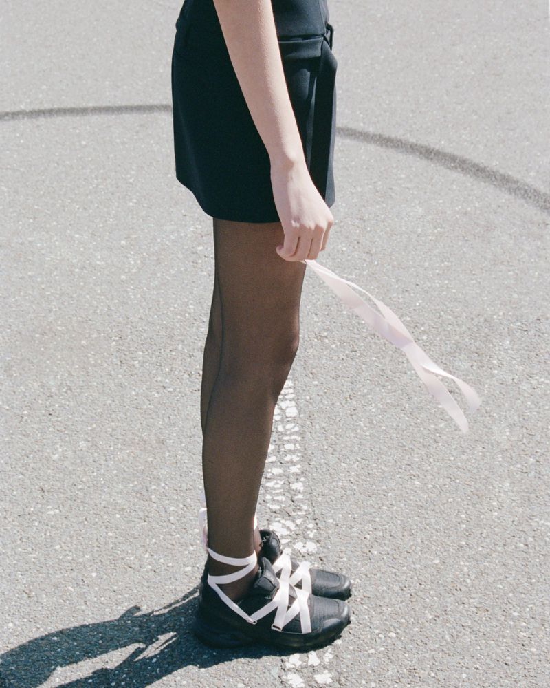 A slender woman in a dark dress, sheer stockings, and lace-up sneakers stands on asphalt, holding a white ribbon, in a medium shot.
