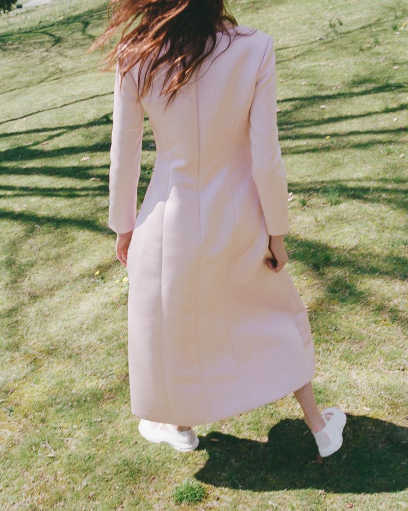 A woman stands on a grassy lawn wearing a long, light pink coat and white sneakers on a bright, sunny day in an outdoor setting.