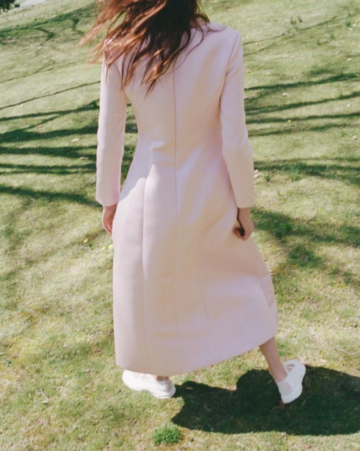 A woman stands on a grassy lawn wearing a long, light pink coat and white sneakers on a bright, sunny day in an outdoor setting.