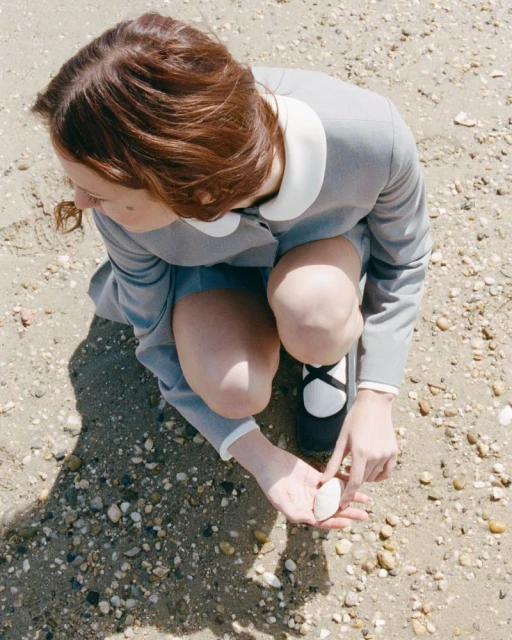 Woman in a gray dress with white collar and black Mary Jane shoes squats on a gravelly beach holding a small white rock.